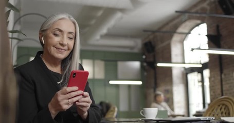 Front view of matured smiling businesswoman using her modern smartphone and looking at screen. Happy modern old woman surfing net and chatting on phone while sitting in cafe.