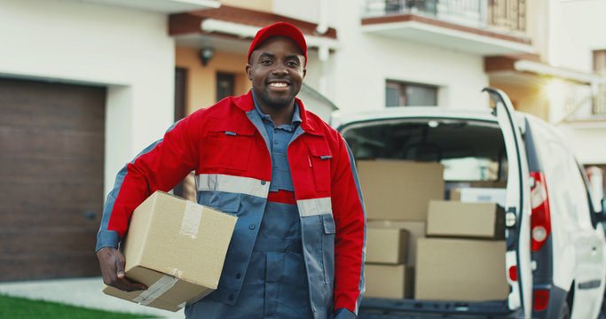 Portrait Of The Young Smiled African American Mailman With A Box Posing To The Camera With Carton Box At The Van. Outdoors.