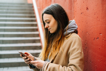 a young girl in a brown coat on the street, standing near a red wall uses her smartphone . street style