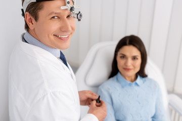 Fototapeta premium selective focus of smiling otolaryngologist in ent headlight holding ear speculum near smiling woman sitting in medical chair