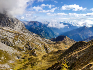 A pathway leading to high peaks in Italian Alps. Sharp slopes on both sides of the valley. Hard to reach mountain peaks. There are many mountain ranges in the back. Serenity and peace. Autumn vibes