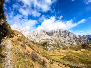 Fototapeta na wymiar Pathway leading to an Alpine cottage in Italian Alps. Sharp slopes on both sides of the valley. Hard to reach mountain peaks. There are many mountain ranges in the back. Serenity and peace. Wandering