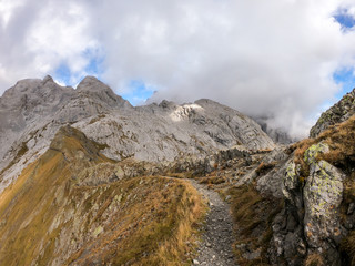 A pathway leading to high peaks in Italian Alps. Sharp slopes on both sides of the valley. Hard to reach mountain peaks. There are many mountain ranges in the back. Serenity and peace. Autumn vibes