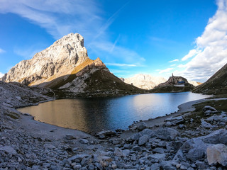 Obraz premium Panoramic shot of a reflection of an Alpine mountain in Wolayer Lake Austria. Completely still surface of the lake. Mountain is catching the first sunbeams. Alpine cottage on the side. High altitude