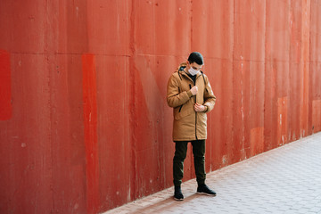 a young man in a brown jacket with a medical mask on his face on the street, near the red wall.