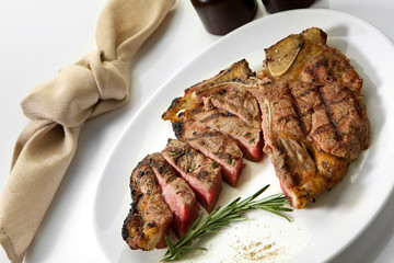 Grilled beef steak with rosemary. Grilled striploin sliced steak. Juicy thick grilled T-bone beef steak seasoned with rosemary fresh of the summer BBQ. Vegetables on a black plate with salt and pepper