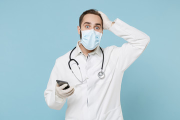 Male on-line doctor man in medical gown sterile face mask gloves isolated on blue background. Epidemic pandemic coronavirus 2019-ncov sars covid-19 flu virus concept Use mobile phone put hand on head.