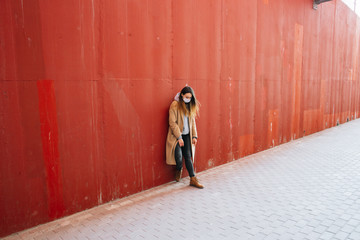 a young girl in a brown coat with a medical mask on her face on the street, near the red wall.