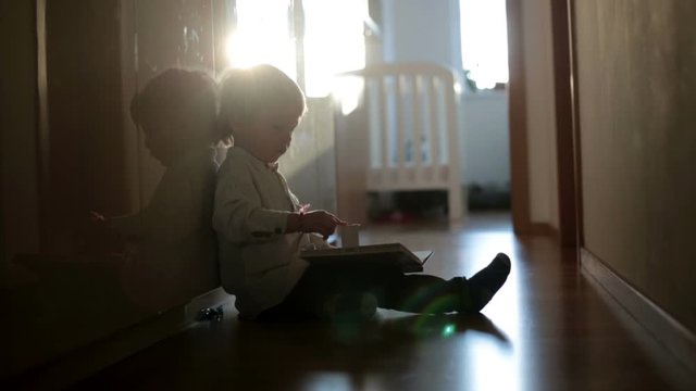 Beautiful toddler blond boy, lying on the floor at home in the hall, reading book, beautiful back light