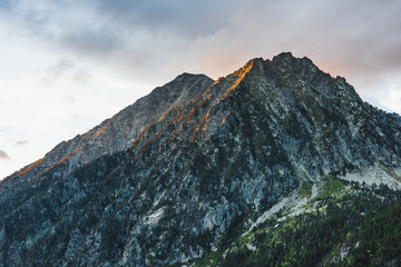 Majestic natural landscape with rocky peak of mountain range covered with green plants against cloudy sky with sunlight