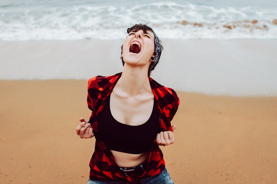 Desperate irritated young hipster female screaming loudly while sitting on sandy beach with sea waves in background