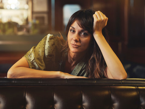 Side view of tired relaxed woman leaning on back of leather black sofa and looking at camera in cafe