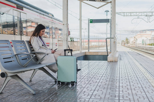 Side view of modern female traveler with suitcase sitting on bench on platform of railway station and using smartphone while waiting for train