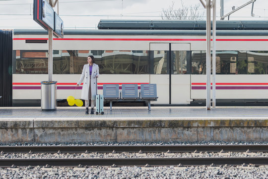 Young female traveler in stylish outfit standing with yellow balloons in hand and suitcase on platform of railway station