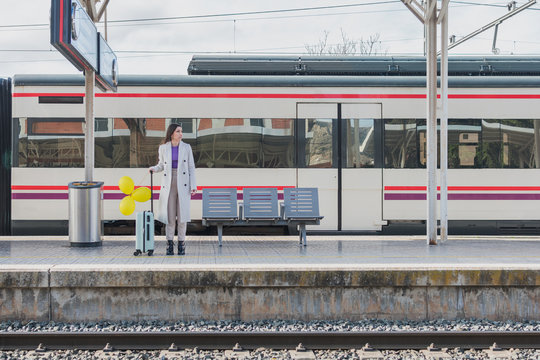 Young female traveler in stylish outfit standing with yellow balloons in hand and suitcase on platform of railway station