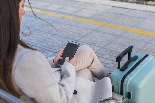 From above copped unrecognizable female traveler with suitcase sitting on bench on platform of railway station and using smartphone while waiting for train