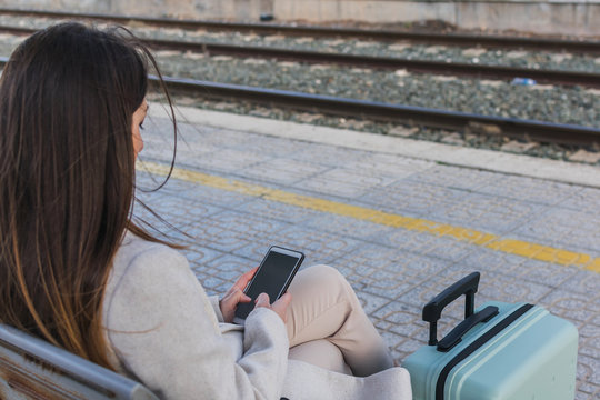 From above copped unrecognizable female traveler with suitcase sitting on bench on platform of railway station and using smartphone while waiting for train