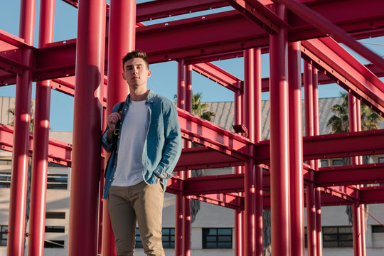 Smart stylish man with backpack looking at camera standing in yard with red metal construction in sunny day