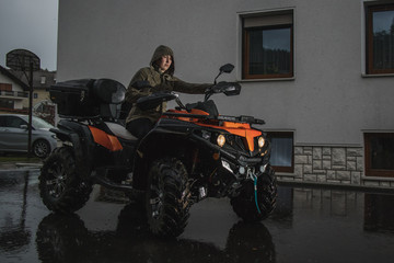 Young caucasian woman is manouvering an orange quad or ATV vehicle while it is raining outside of the house.