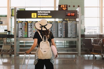 Back view of woman in hat with suitcase and pillow looking at schedule in airport