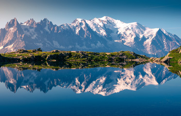 Range of white snowy peaks and hilly seaside reflecting in clear motionless lake in sunny day in Chamonix, Mont-Blanc