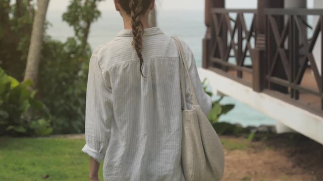 woman walks near rest hotel against tropical plants and storming ocean backside view slow motion. Concept exotic travel environment