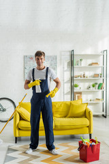 Fototapeta na wymiar happy cleaner in overalls holding rag and spray bottle in living room