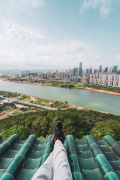 Crop legs of man chilling on roof with view of contemporary Nanning cityscape and green shore of river below, China