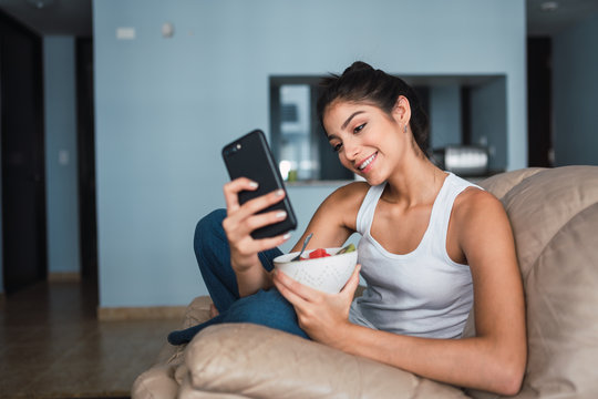 Beautiful brunette Hispanic woman holding fork with slice of fruit and sitting on settee with bowl of kiwi and watermelon and browsing in mobile phone