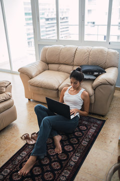 From above beautiful brunette Hispanic woman typing on laptop and sitting on floor on little carpet near settee and window