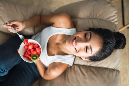Beautiful cheerful brunette Hispanic woman holding fork and bowl of kiwi and watermelon and sitting on settee