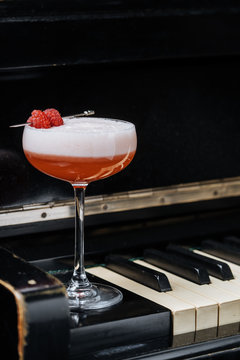 Red alcohol cocktail in stylish glass with white foam decorated with fresh raspberry on piano keys in restaurant