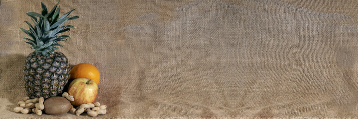 Horizontal banner. Pineapple orange apple and peanuts on raw yuta canvas background.