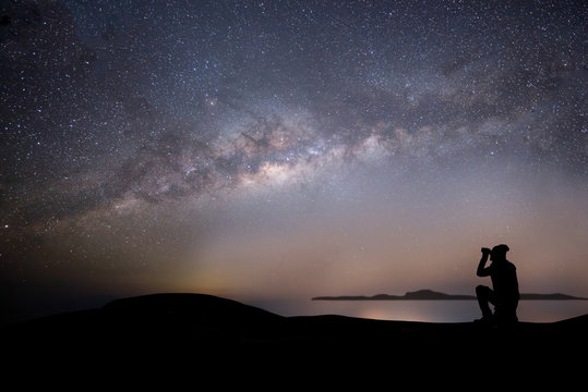 Milky way night panorama with silhuettes of trees and a man intent on photographing the sky.