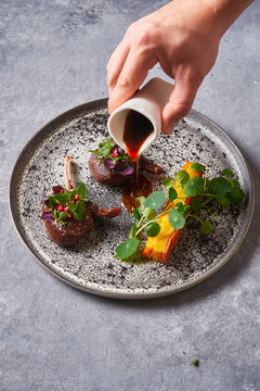 From above cropped unrecognizable person adding sauce to tasty meat medallions with herbs on plate on gray stucco table