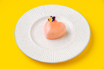 From above small heart shaped dessert with pink icing and small flower placed on plate on yellow background