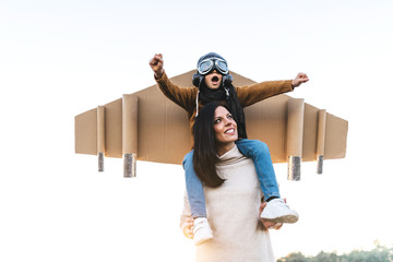 Happy cute boy wearing goggles and cardboard wings while sitting on mother shoulders and imitating aviator on meadow in backlit
