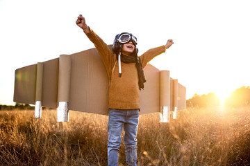 Happy kid in goggles and cardboard wings raising hands during game on field in backlit