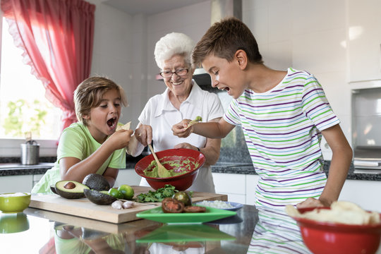 Cheerful grandma and boys tasting homemade guacamole paste with tortilla chips in kitchen