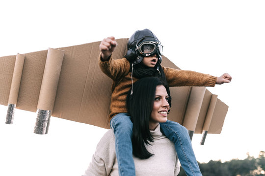 Happy cute boy wearing goggles and cardboard wings while sitting on mother shoulders and imitating aviator on meadow in backlit