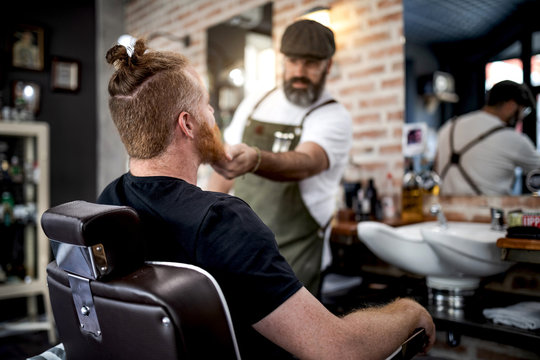 Barber trimming beard of redhead man sitting in barbershop