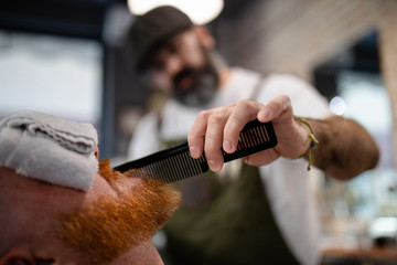 Blurred unrecognizable barber with comb fixing redhead man beard with towel covering eyes sitting in barbershop