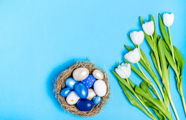 Easter eggs of blue flowers in a decorative nest of straw and a bouquet of white tulips on a blue...
