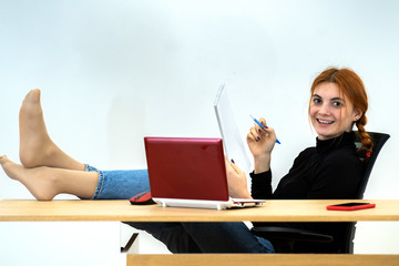 Happy young office worker woman sitting relaxed with feet on table behind working desk with laptop...