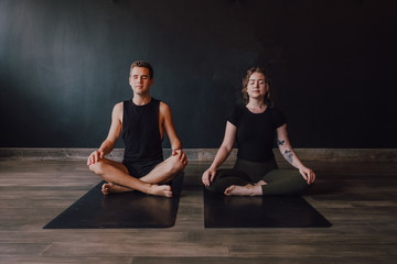 Calm young woman and man in sportswear with eyes closed and legs crossed meditating together while sitting in padmasana position against back wall in contemporary yoga studio