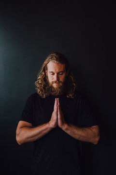 Handsome male athlete in sportswear with namaste praying hands performing yoga kakasana against black wall of contemporary workout room