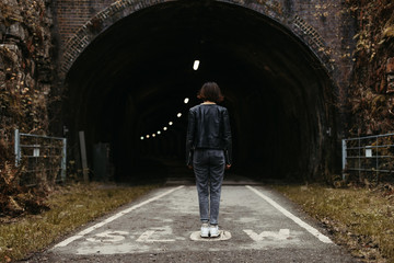 Back view of stylish woman in black leather jacket looking at dark tunnel with illumination inside on marked with inscription slow road in United Kingdom