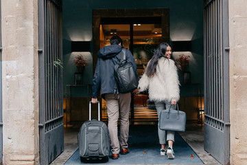 Back view of happy stylish young couple in love with suitcase and bags holding hands and entering modern city hotel during travel while woman looking at camera over shoulder