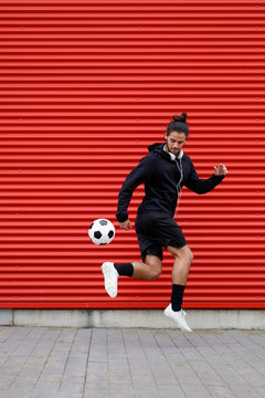 Side view of young professional male soccer player in black sportswear making feint against red roller shutter door on street