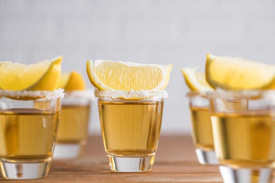 Row of glass sots with golden tequila and slices of lemon on wooden table with white wall on blurred background